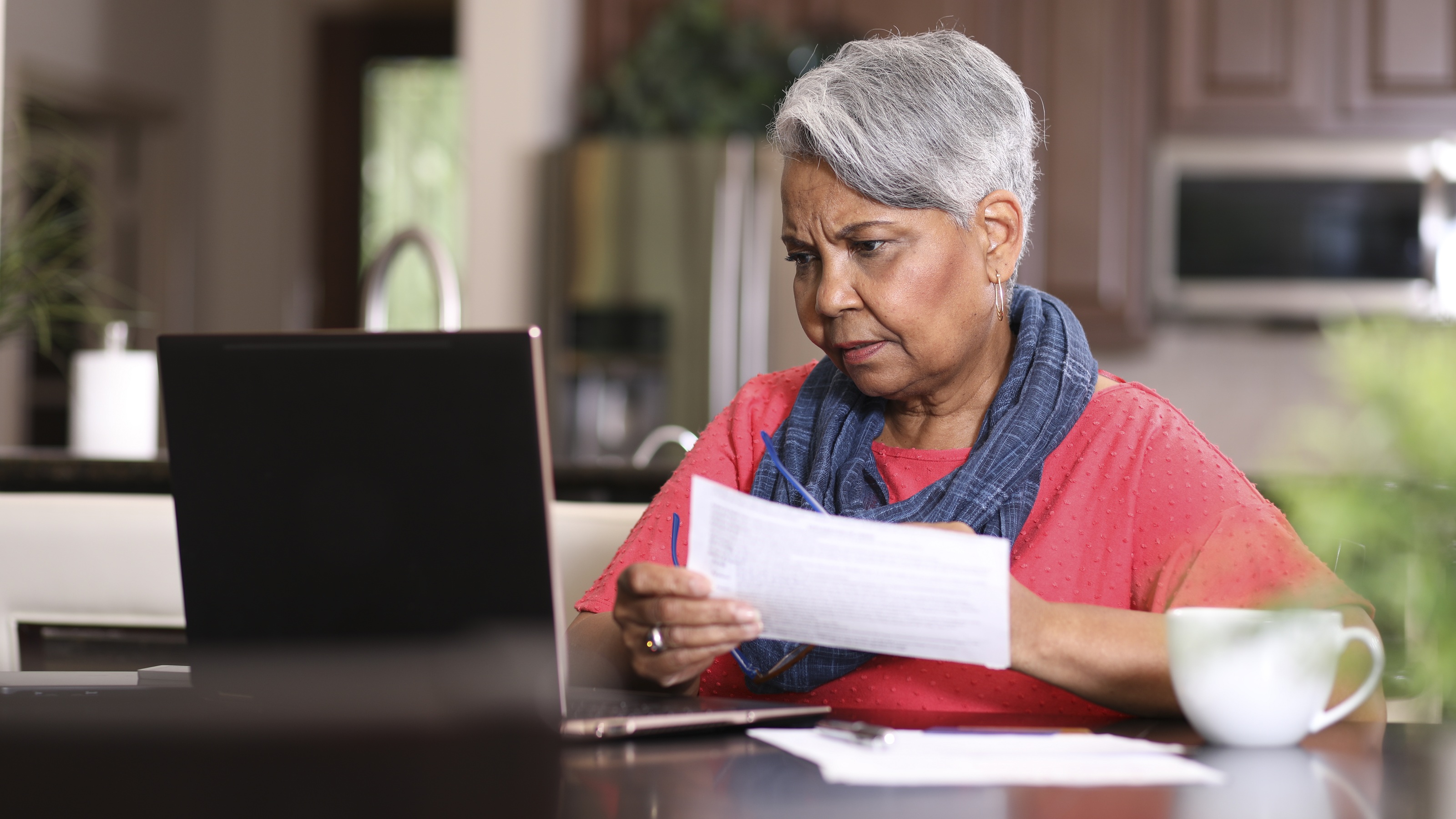 An older woman looks concerned as she looks over paperwork with her laptop open on her dining room table.