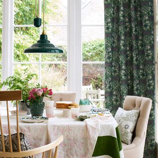 A dining room with dark green pendant light and curtains and beige padded dining room chairs