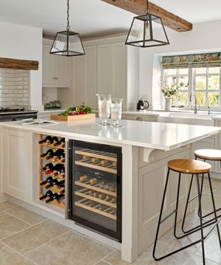 A white kitchen island with a wooden wine rack and black wine fridge on the side of it, two glasses and a box of vegetables on top of it, two black glass pendant lights above, and two wooden stools to the right