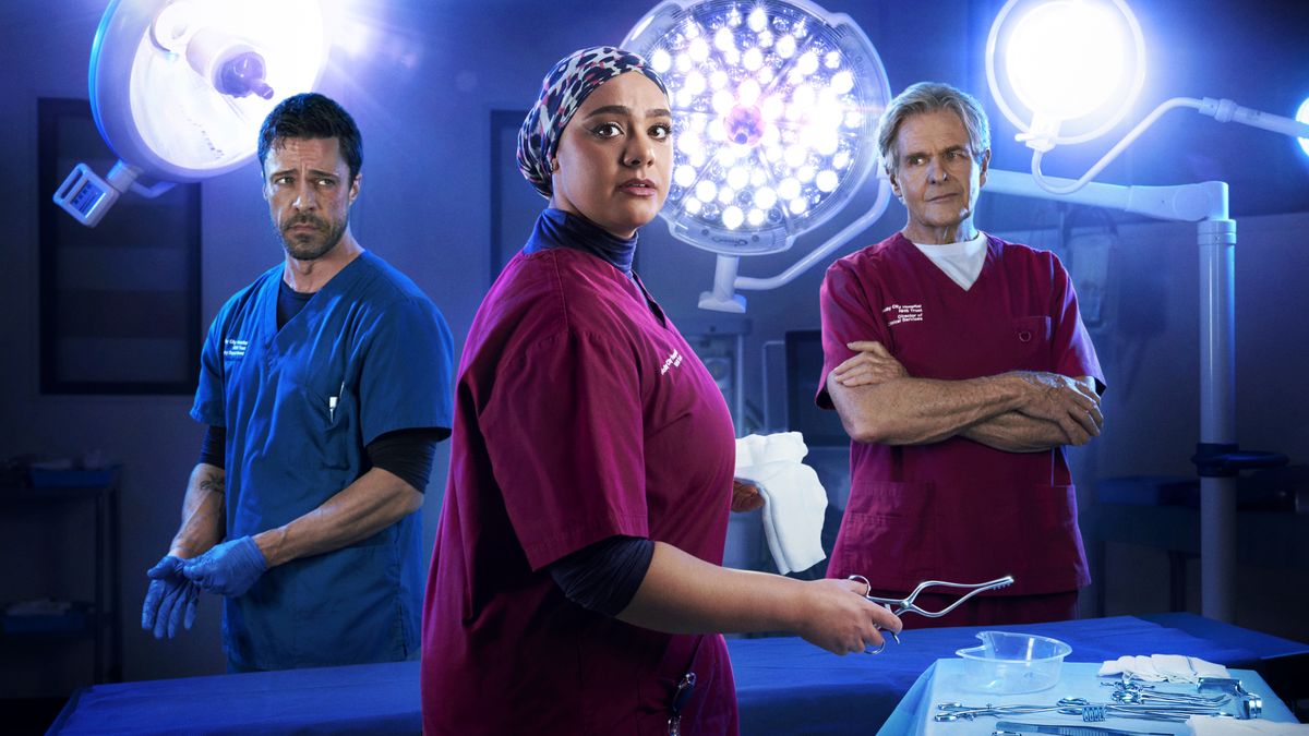 New promotional image for Casualty&#039;s 12-parter Internal affairs. It shows Rida front and centre dressed in maroon surgical scrubs with a printed headscarf and holding a pair of surgical forceps, looking serious and at the camera. Behind her are Flynn in blue scrubs and blue gloves and Russell in maroon scrubs with his arms crossed. The backdrop is a blue surgery with bright surgical lights. 