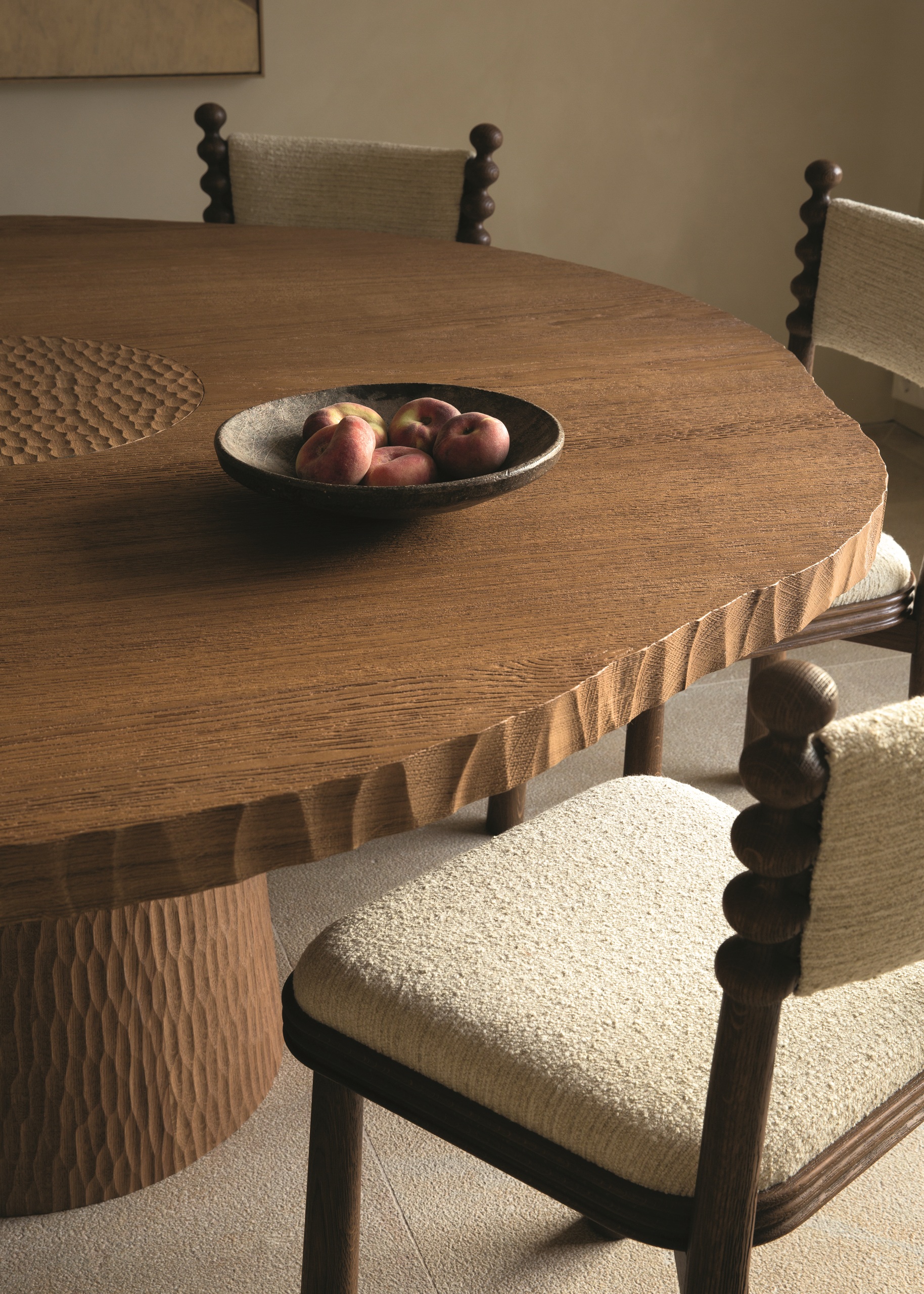 a close up of a round timber dining table with textured edge and hand-gouged pedestal base, with cream floor and cream boucle dining chairs with bobbin details