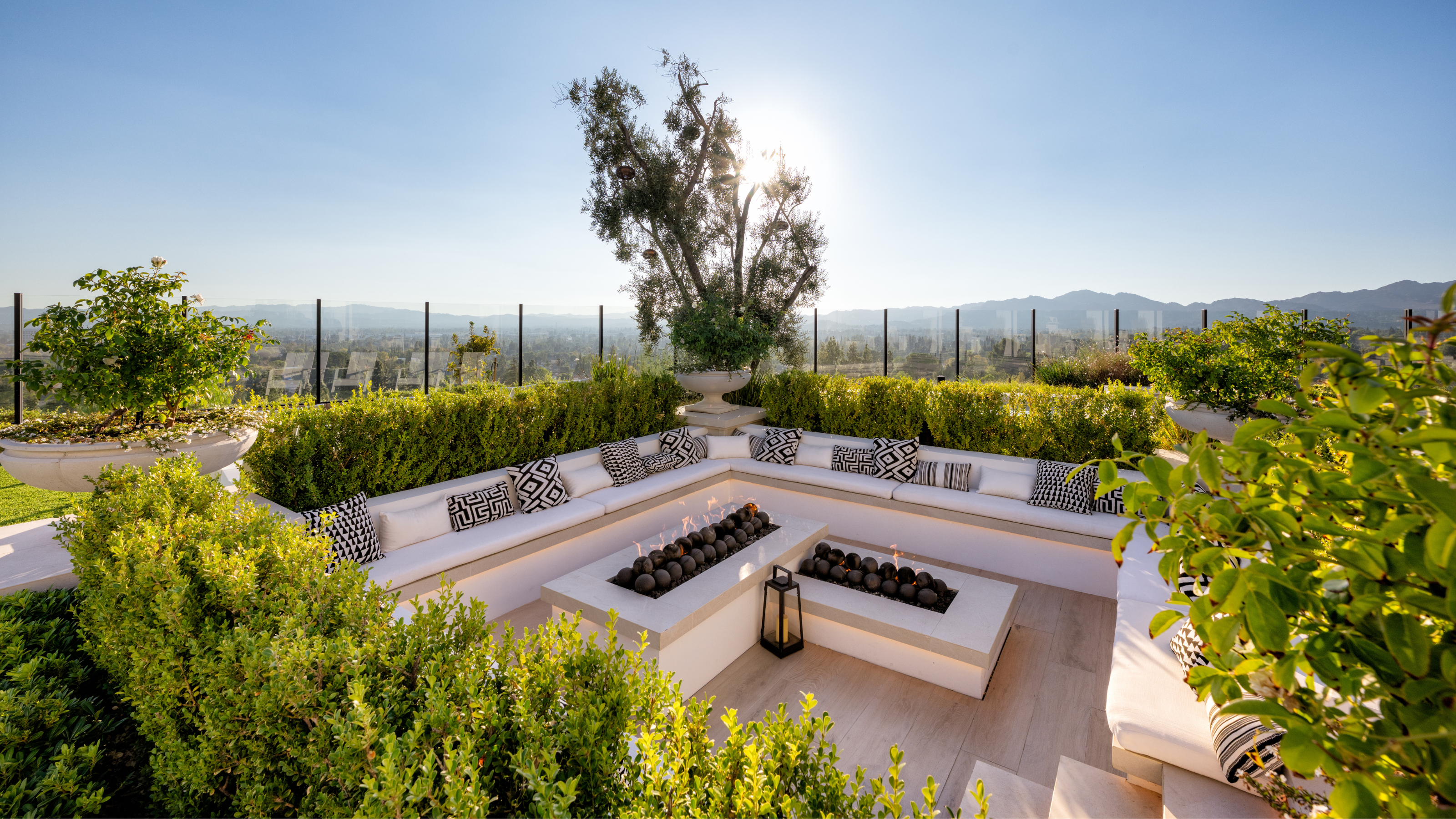 A modern built-in outdoor seating area with two fire pits and a lantern, surrounded by greenery and a clear blue sky with the sun just out of view behind a tree with the fence along the perimeter
