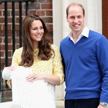 Princess Kate stands outside the hospital holding Princess Charlotte who is wrapped in a white blanket, while Kate wears a yellow floral dress and Prince William wears a blue sweater