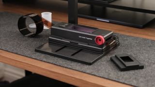A black film scanning stand setup on a desk, featuring a base with a built-in unlit panel, a vertical mounting column, and a strip of 35mm film feeding through the holder.
