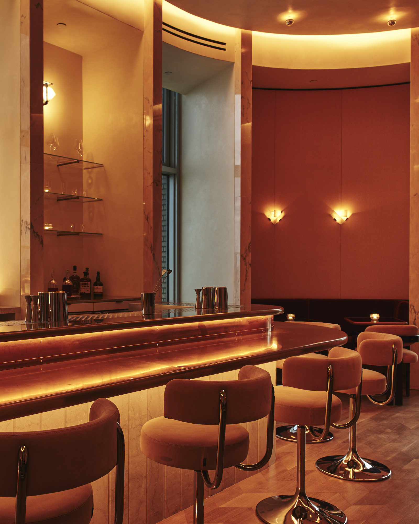 A dimly lit, panoramic bar decorated with 1980s-style chrome and orange velvet chairs, golden-lit sconces, and sleek, back-lit surfaces overlooks a city at dusk.