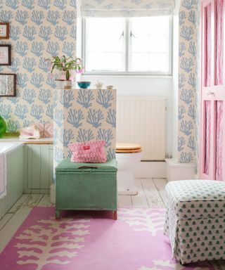A bathroom with white and blue coral patterned wallpaper, a window, pink door, seafoam green stool, a bright pink coral rug, and white wooden distressed flooring