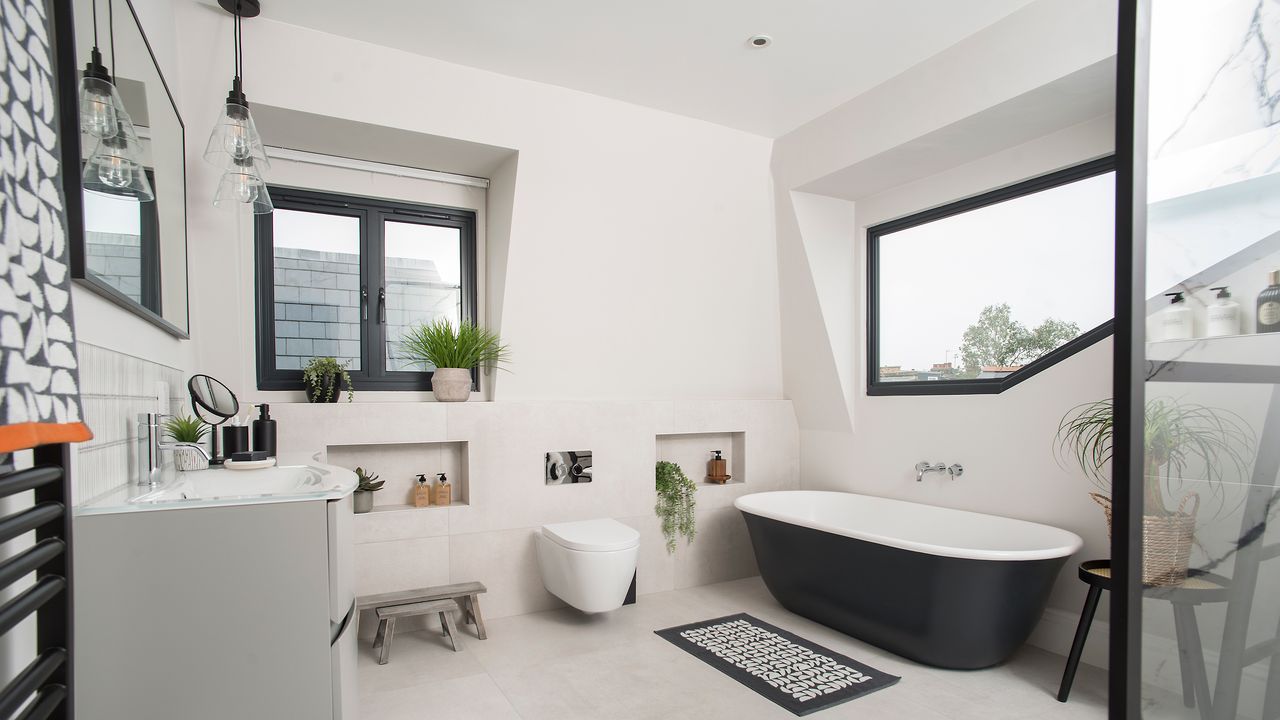 A modern loft bathroom with black bath, black-framed windowsill, white toilet and vanity and duo of clear pendant light fixtures