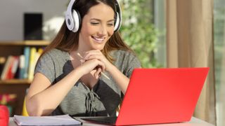 A woman sitting at a table in a library using a laptop with a red cover and she is wearing headphones