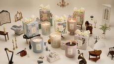 White background with five pastel candles and their packaging boxes arranged around miniature figurines of antique furniture.