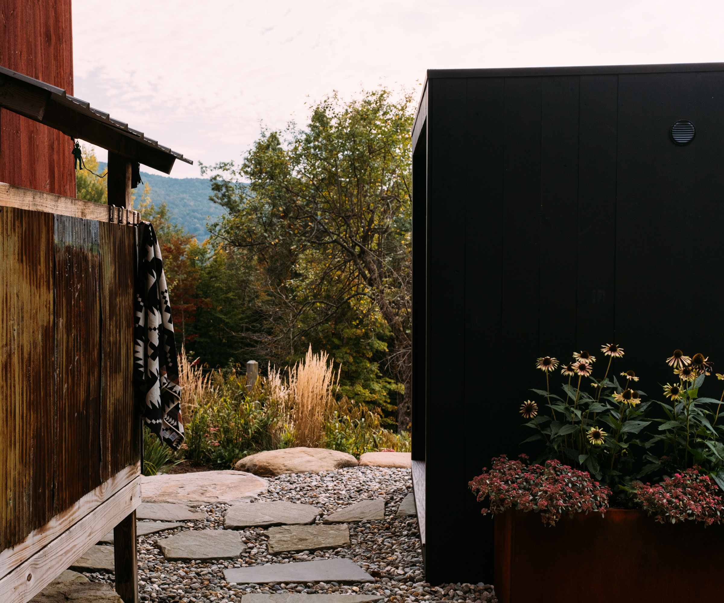 gravel and flagstone path with ornamental grasses, flowers and trees, and a black painted shed plus timbered building on stilts