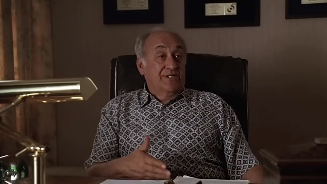 As Hesh in The Sopranos, Jerry Adler, who is deceased, is sitting at a desk with awards displayed behind him.