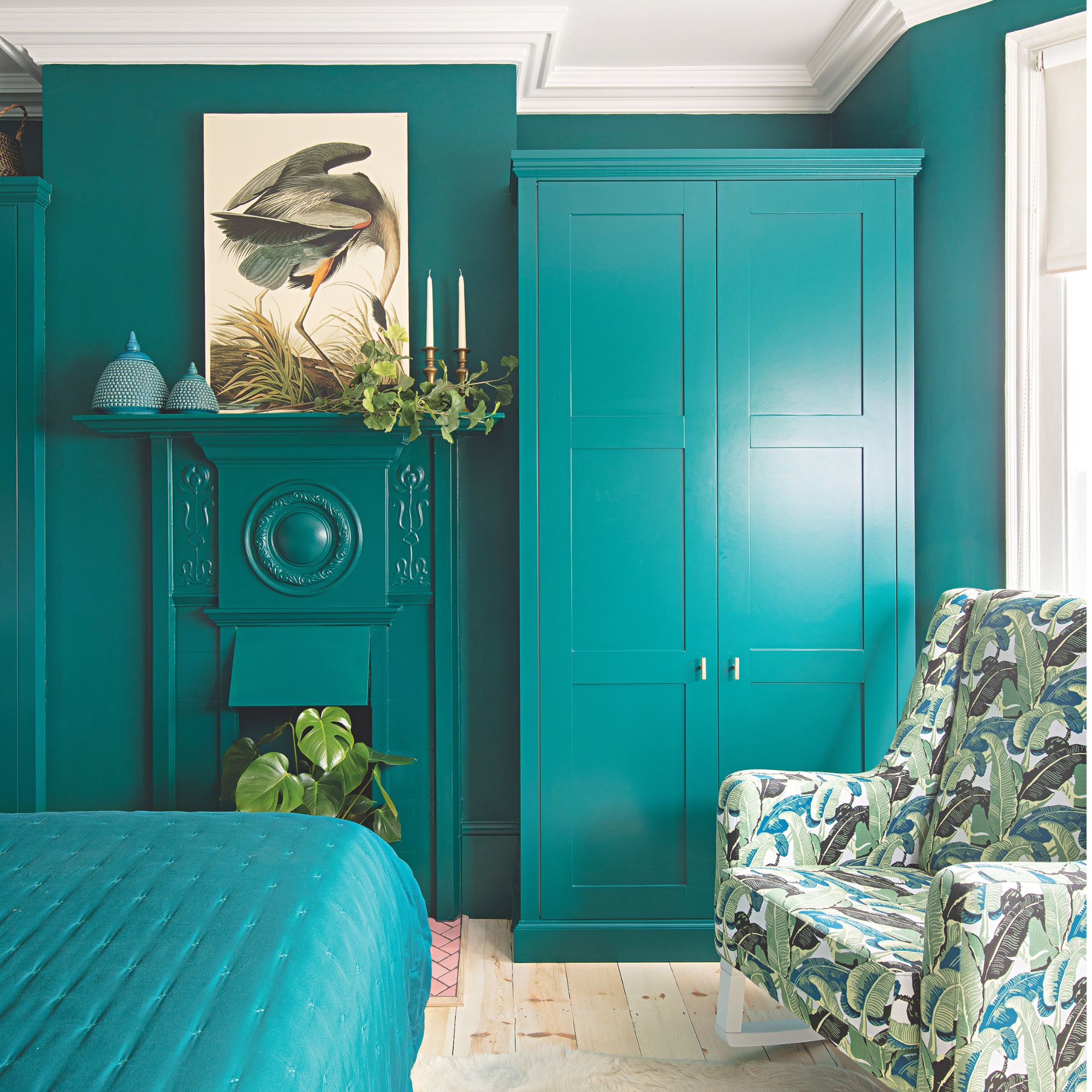 Teal colour drenched bedroom with teal painted walls, freestanding wardrobe, fireplace and bedding, with a tropical leaf print accent chair on wooden flooring