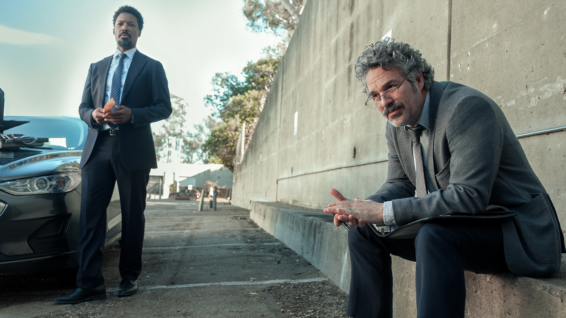 Detective Tillman standing next to a car as Detective Lubesnick sits on a wall nearby in Crime 101