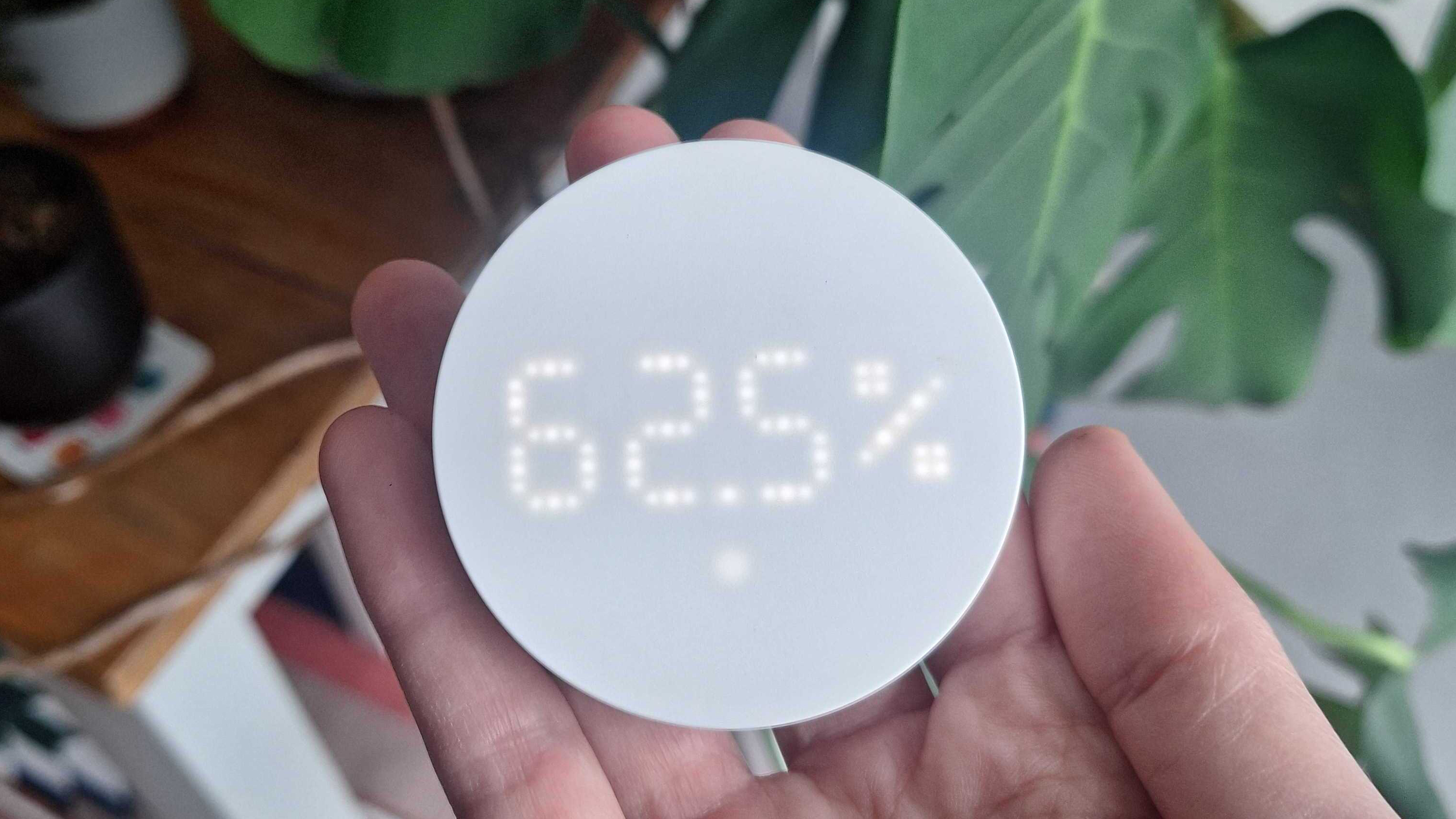 IKEA Timmerflotte temperature and humidity sensor in writer's hand, showing a 62.5 per cent humidity reading on screen. With a wooden table and plants in the background