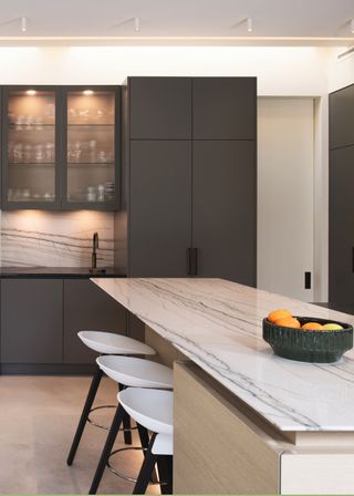 Dark tones fluted kitchen cabinets with a large cupboard next to it. There is also a large marble island in the kitchen with a fruit bowl on top.