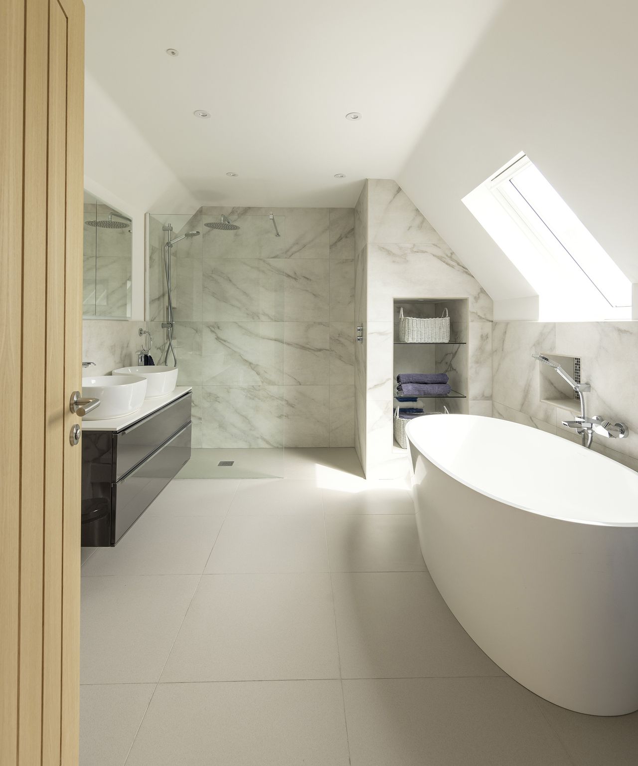 A large bathroom with light coloured flooring and marble effect panels. There are his and hers sinks and a roof light