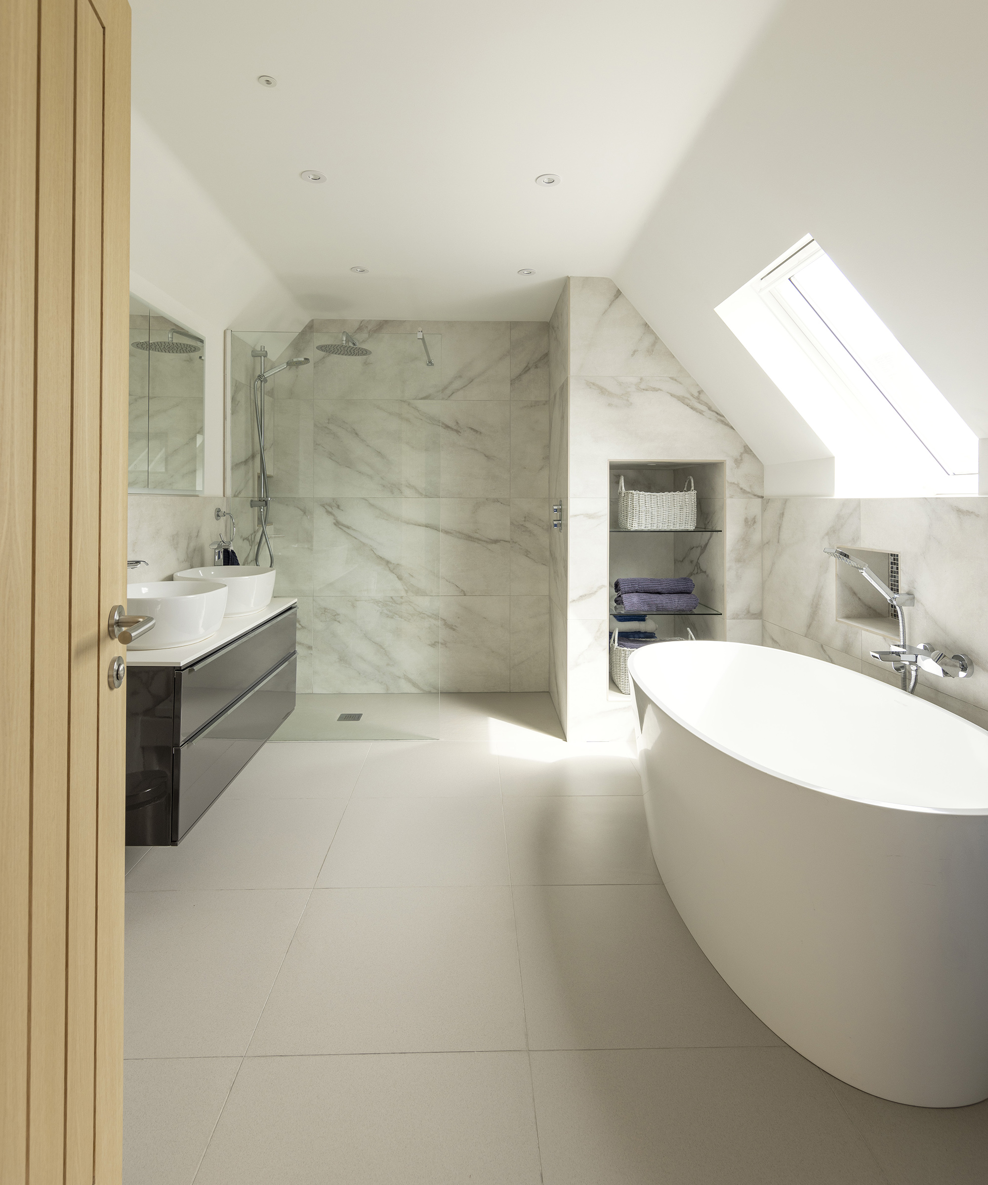 A large bathroom with light coloured flooring and marble effect panels. There are his and hers sinks and a roof light
