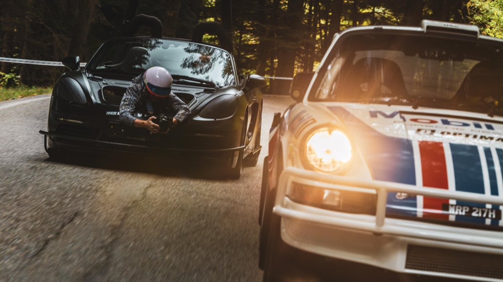 A photographer in a black Porsche camera car, low to the ground, shoots a classic white rally-style Porsche with its bright headlight illuminated on a forest road.