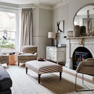 a neutral living room with a decorative mantle around a woodburner fireplace, with large window with full length neutral curtains, paisley armchairs and a brown and white stripe footstool