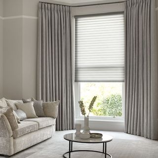 a grey and white living room with grey curtains at a bay window, with light carpet and a white velvet sofa with matching cushions