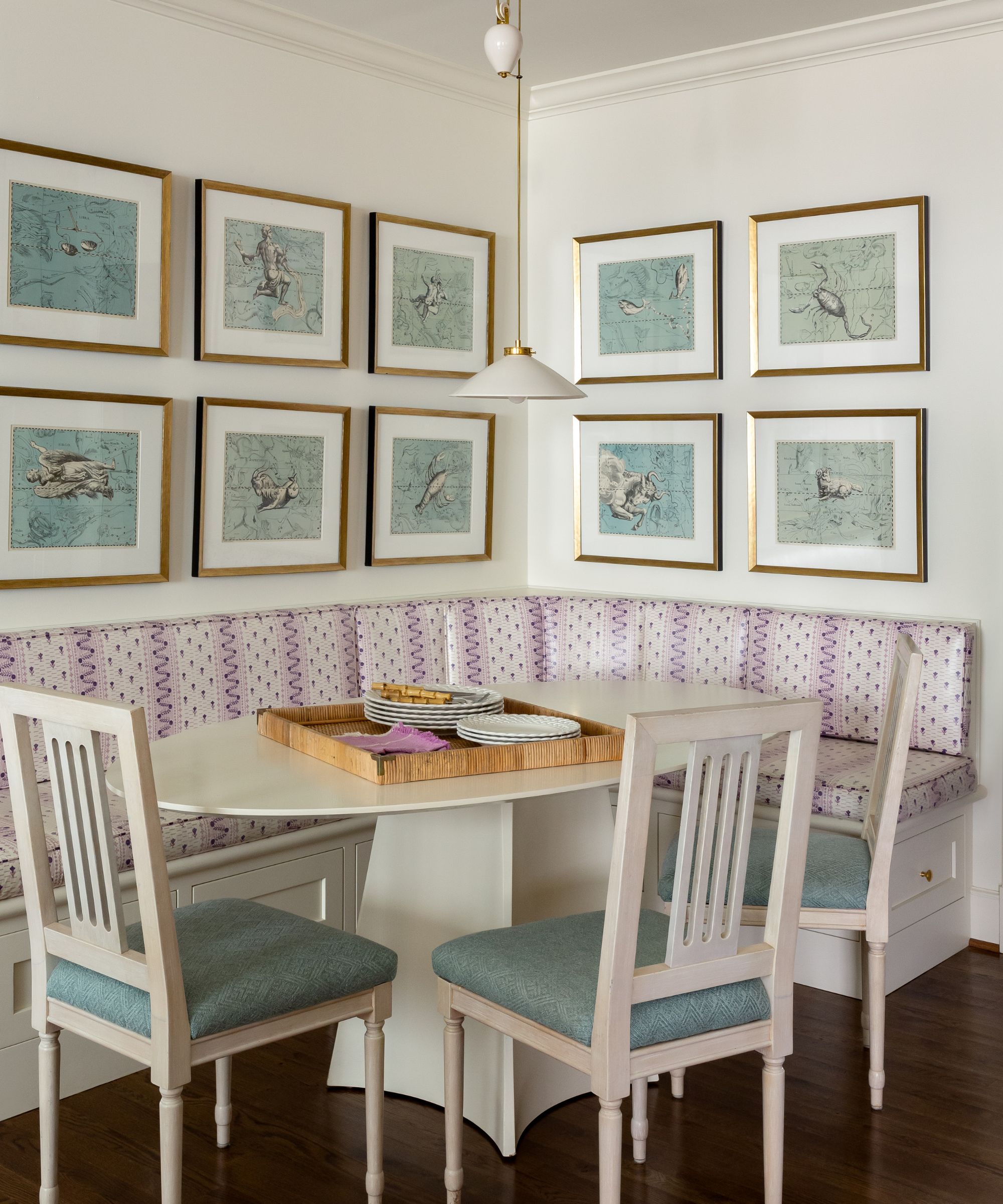 A breakfast nook with white walls, a gallery wall with blue artwork in matching frames, lilac banquette seating with a white oval table and three chairs with blue seat cushions.