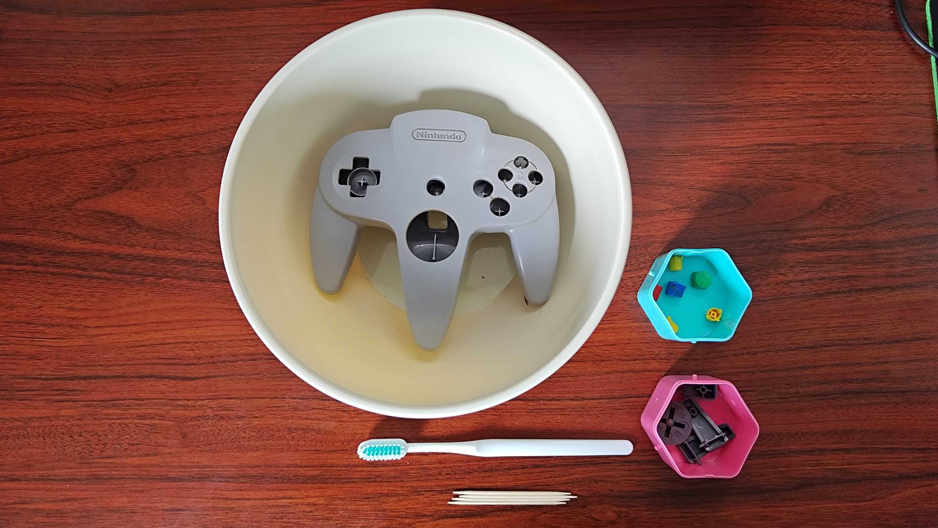 N64 controller shell in plastic basin with buttons in a blue and pink hexagon container next to white toothbrush and toothpicks on woodgrain desk.