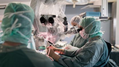 A look inside a surgery room, with various people wearing blue scrubs and face masks and gloves, holding tools above a patient on a table