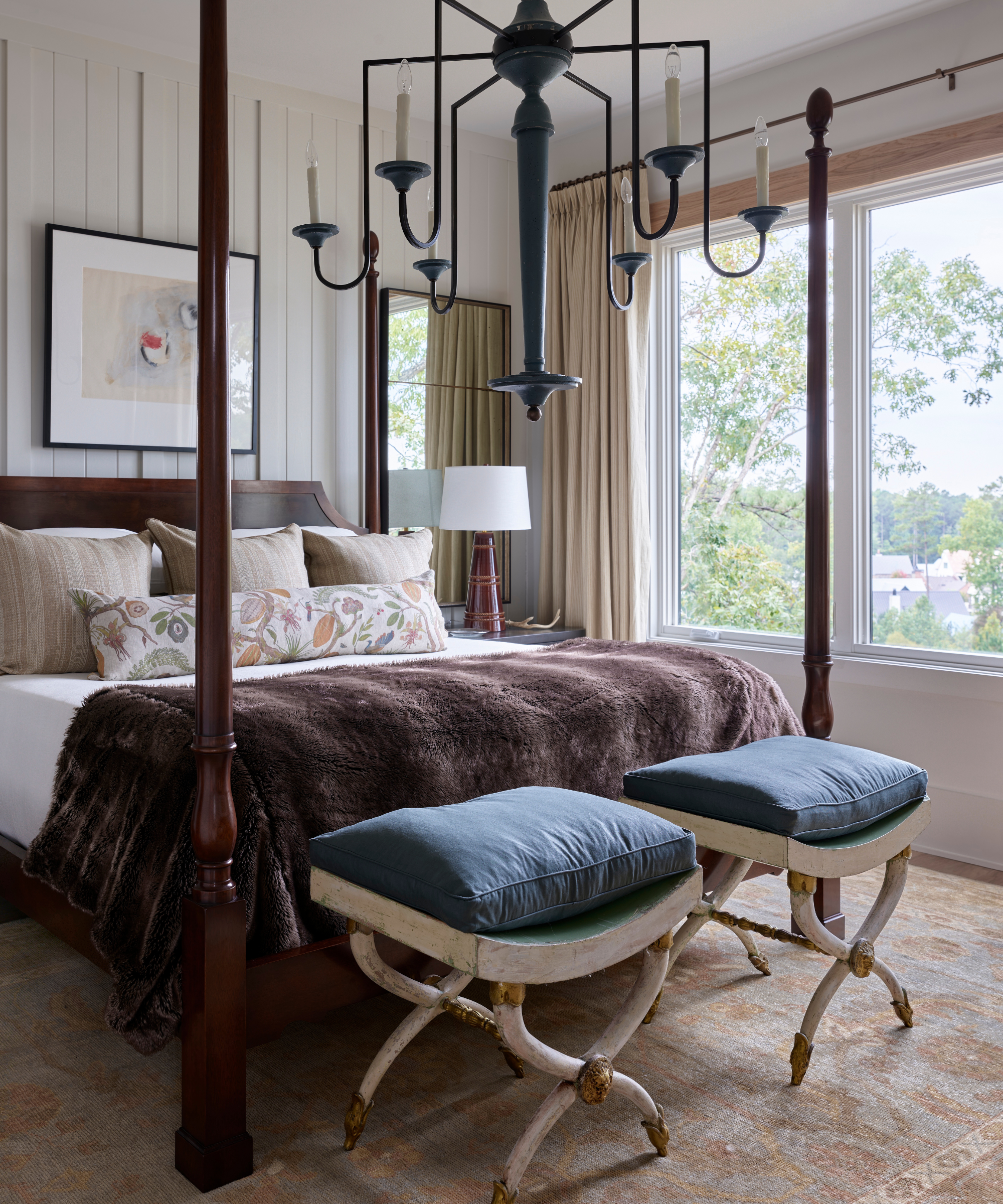 a vintage-inspired bedroom with a four-poster bed in dark wood, two vintage stools at the foot of the bed, a large candelabra chandelier, and a light-washed persian rug on the floor