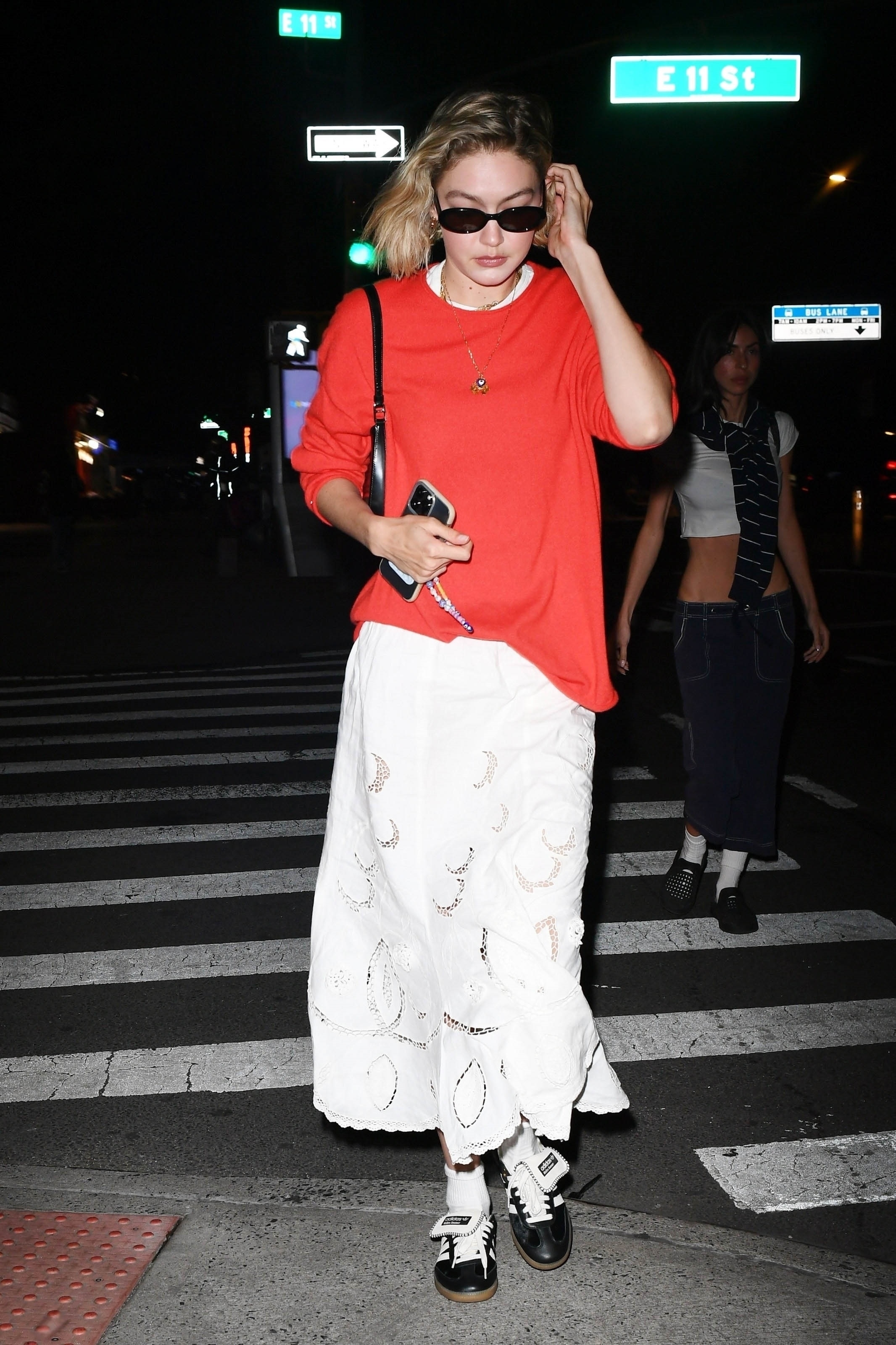 Gigi Hadid walking in New York City wearing a red T-shirt, sunglasses, sneakers, and a white eyelet skirt.