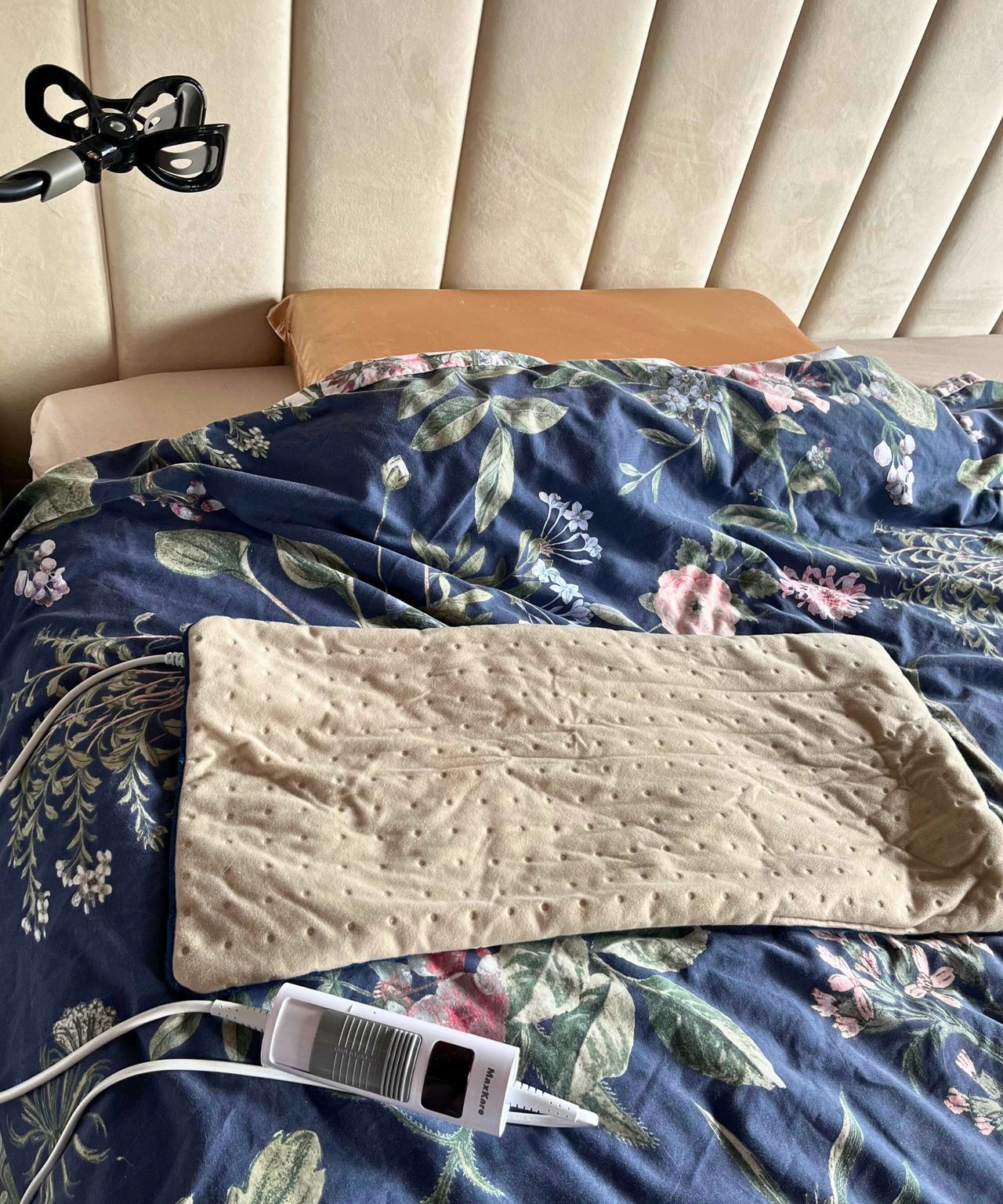 A cream heating pad on my bed which has a dark blue floral comforter and paneled upholstered cream tall headboard