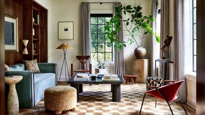 A living space with tall windows dressed with striped curtains, and a recessed, dark wood feature wall to the left. In the centre of the space is a square, stone coffee table with a blue sofa and beige textured footstool, as well as a red modern armchair to the right.