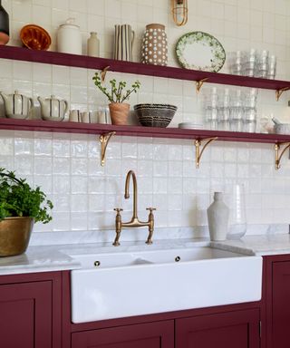 A burgundy kitchen sink unit with a white basin and gold taps with two long burgundy shelves above it with plates, vases, glasses and plants on top of them