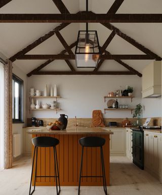 A kitchen with dark brown wooden beams above it, a black iron pendant light, and a kitchen island with orange panels and two black seats in front of it