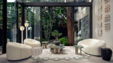 A beautiful living room with glass windows looking out onto a lush backyard, white sunken couches and sofa seaters, a clustered coffee table and side tables, a geometric rug, a standing paper lantern lamp, and a pair of floor lamps