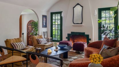 A white living room with colorful decor in the form of mustard sofa seaters with striped pillows, green window trim, a green border tile fireplace, deep burgundy poufs, a cinnamon orange sofa, wood chairs with black frames, a low wood coffee table an da checkerboard rug with potted plants
