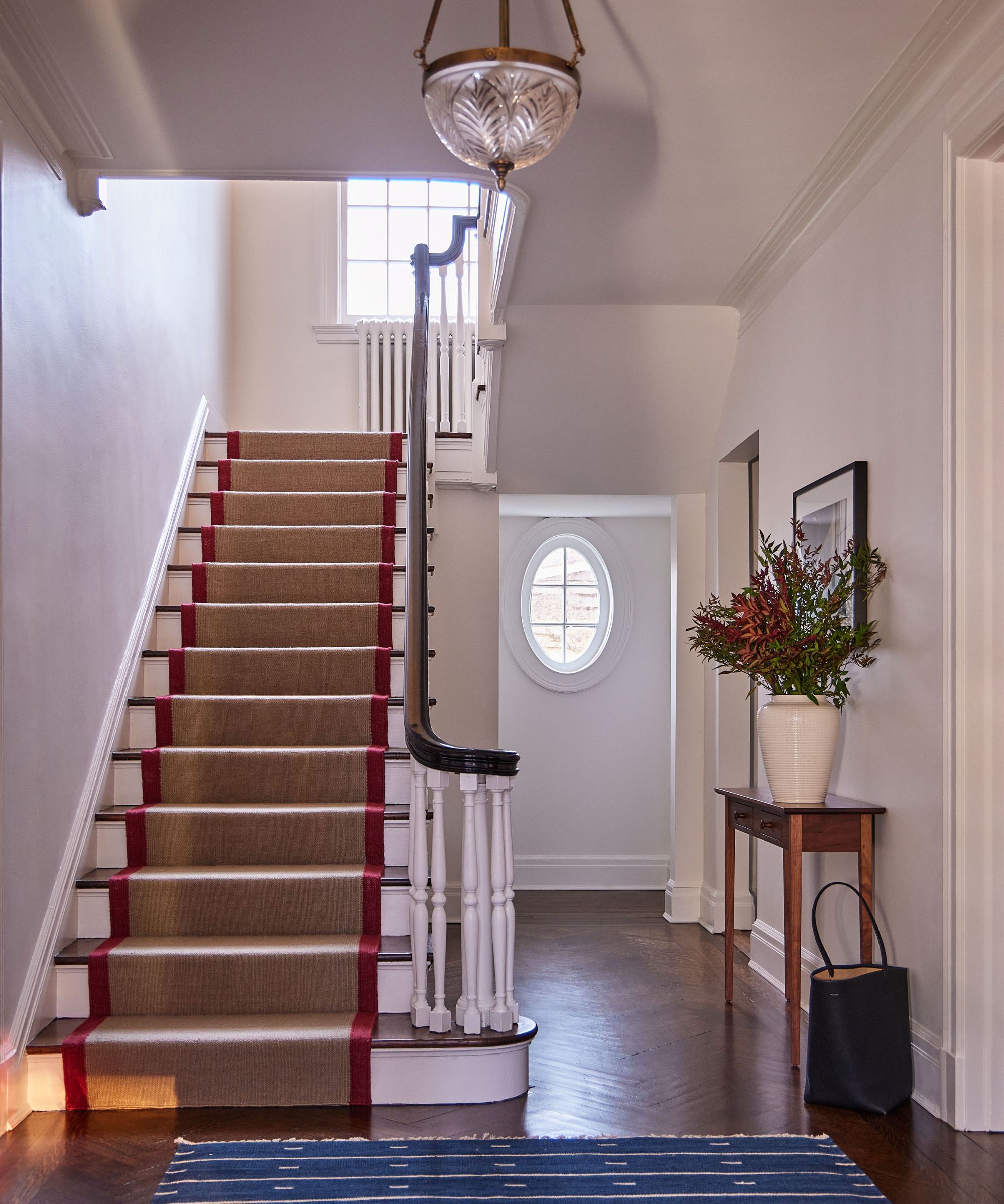hallway with white walls, dark wood floor, blue rug, staircase with jute runner and glass vintage pendant light