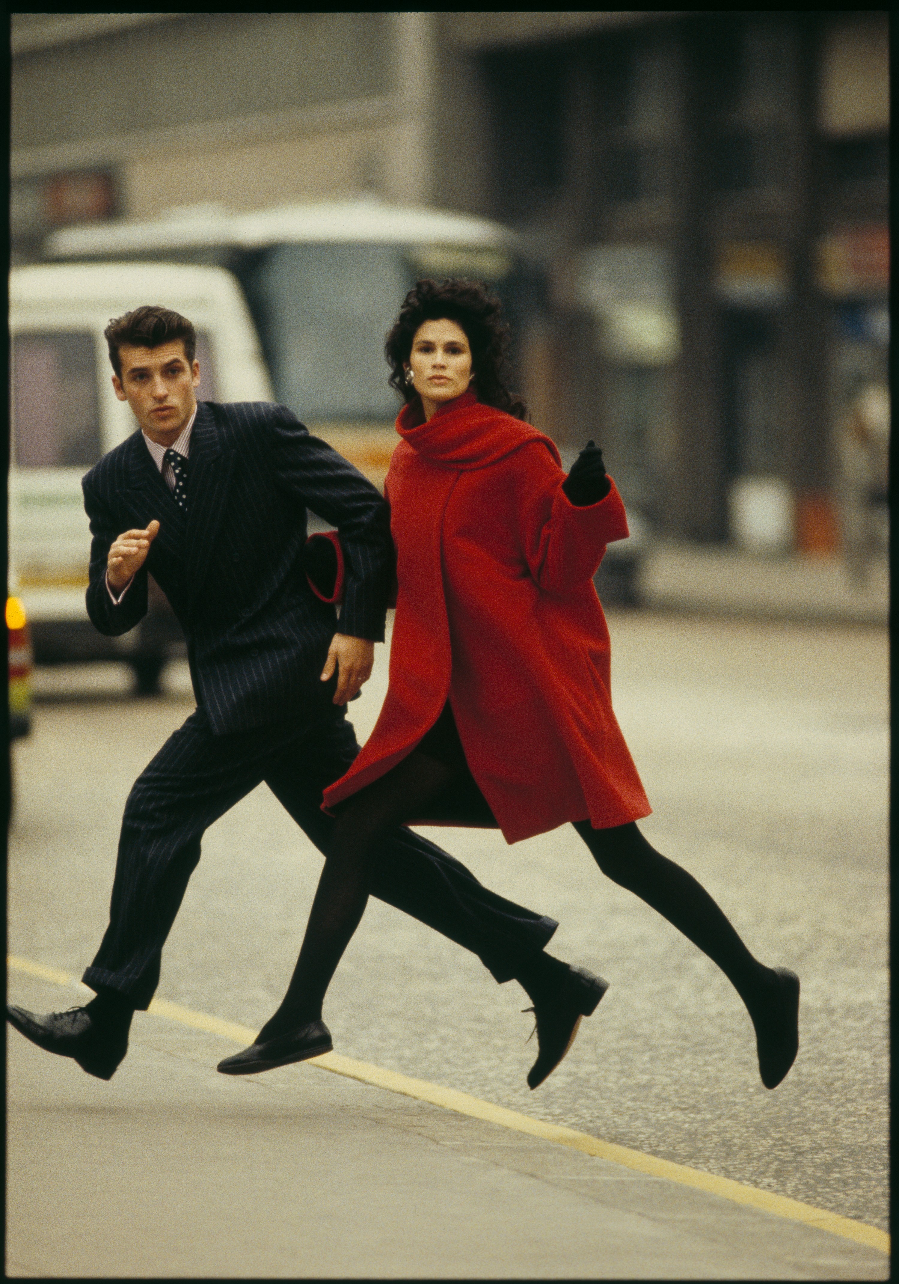 A man in a pinstripe suit and a woman in a vivid red coat leap together across a city street, both fully airborne.