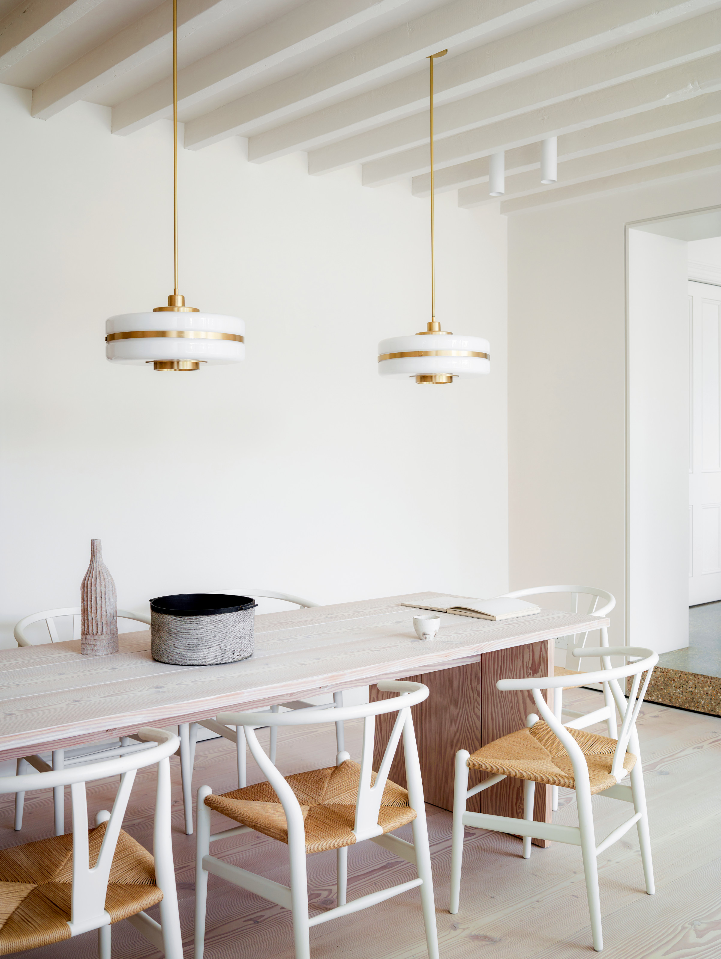 White dining room with pale wood table, white wood and rattan chairs and white and brass pendant lights