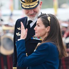 Queen Letizia wearing a blue suit and covering her face with her hand as King Felipe looks down on her 