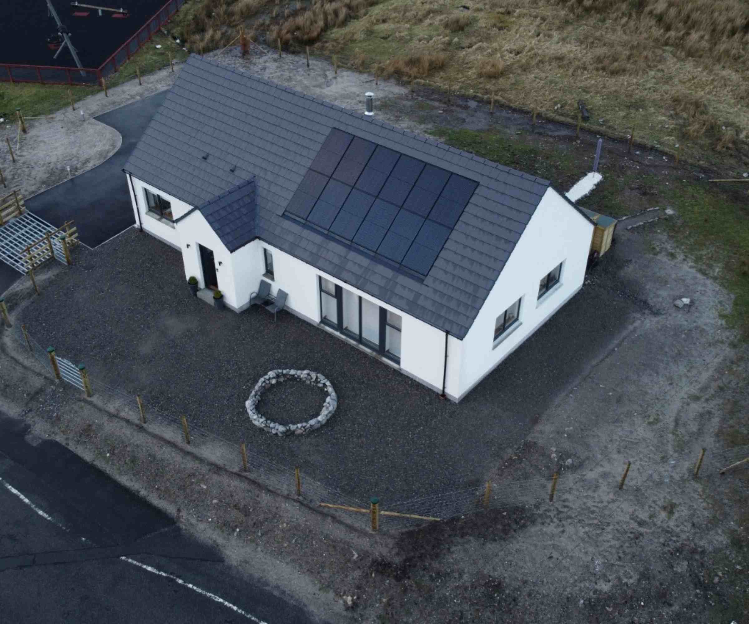 An aerial shot of a single-storey white-rendered new home, with solar panels installed on the slate roof. The new house, built in timber frame from company Norscot, sits on a small site, due to be landscaped, in Scotland.