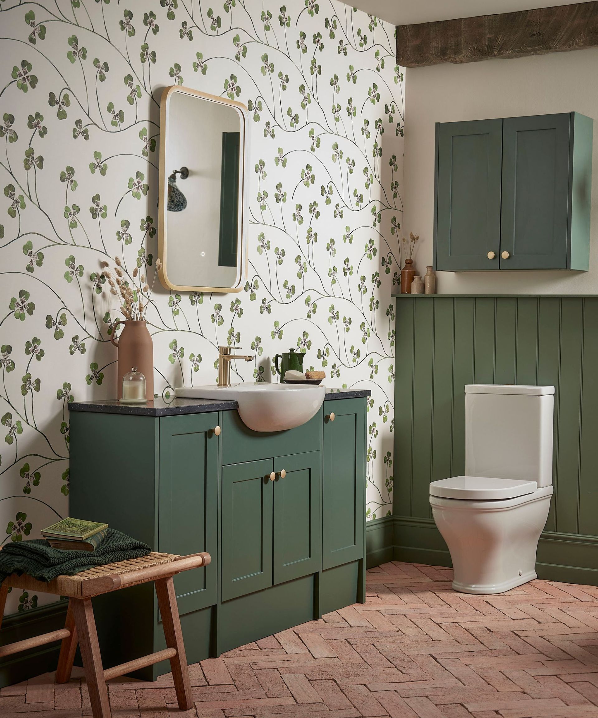 A green bathroom with white clover wallpaper, a gold curved mirror, a green sink unit, brick flooring, and a white toilet