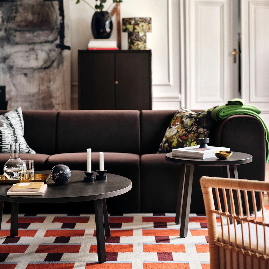Image of a living room with a dark brown sofa in the center and two round, black coffee tables in front of the sofa. There is an orange and white rug with a woven pattern on the ground. The coffee table has decorative objects like candles and vases.