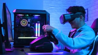 A gamer wears a VR headset and iss teering a plastic wheel at their desk while sat in front of their RGB PC gaming setup