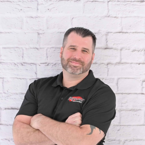 Image of a man in a black collared shirt standing with his arms folded in front of a white brick wall. 