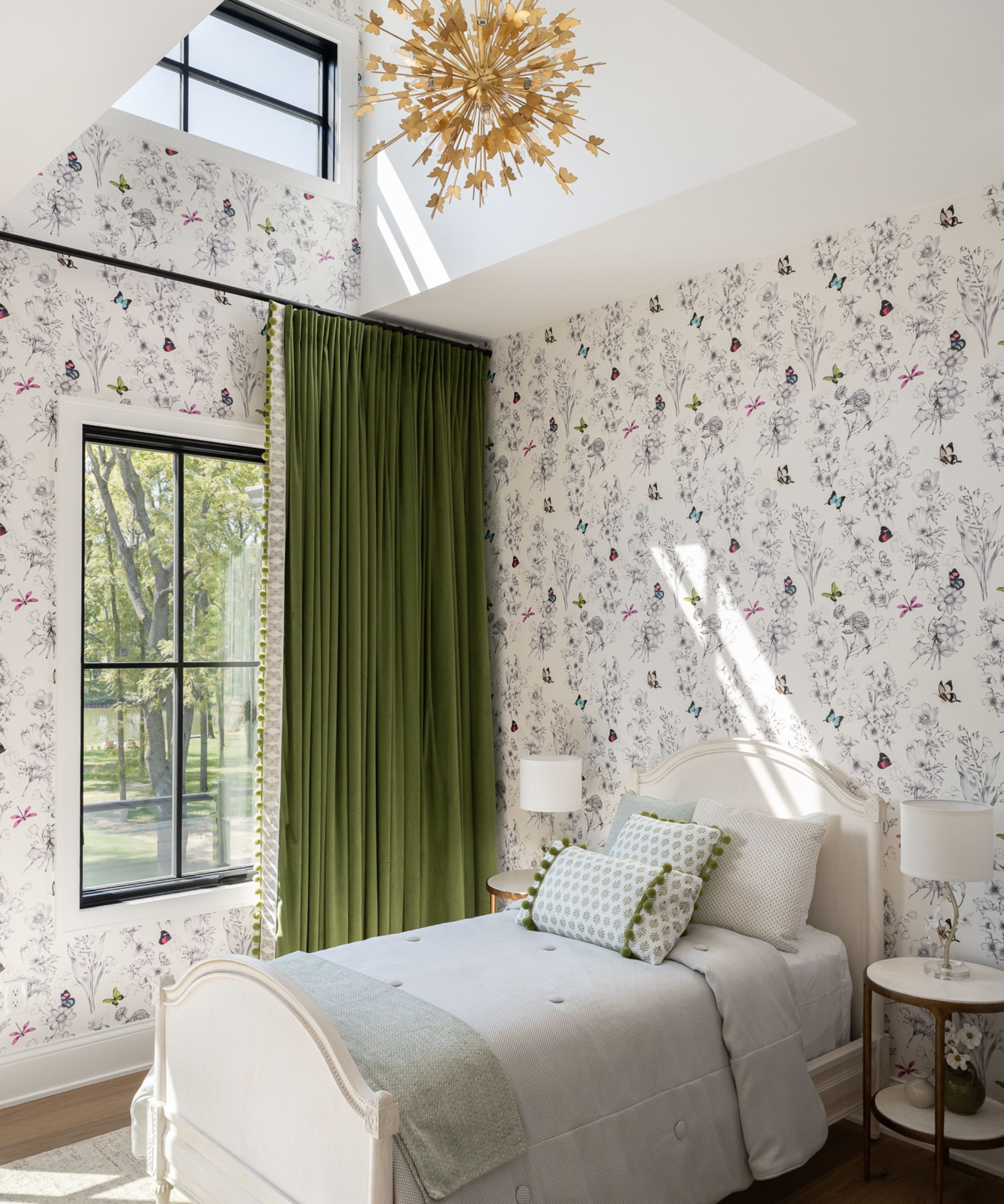 A whimsical child's bedroom with butterfly-patterned wallpaper, a white bed with green pom-pom trim pillows, vibrant green velvet curtains, and a gold starburst chandelier.