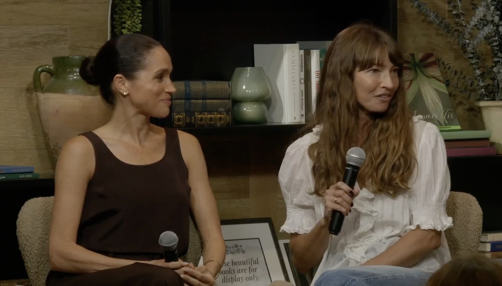 Meghan Markle in a brown outfit sitting next to her friend Courtney Adamo, holding a mic