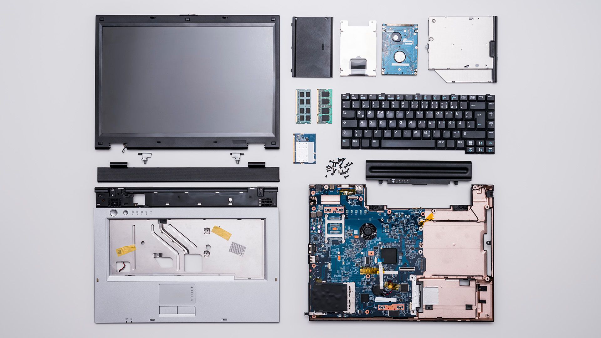 A disassembled laptop, with motherboard, keyboard, processor, disk drive, memory, display, and other parts visible, placed on a white surface.