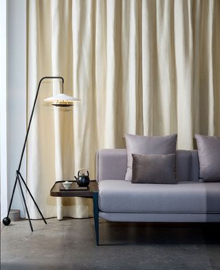 The Float sofa by Michele De Lucchi for Stellarworks photographed against the backdrop of a white curtain, and with a floor lamp next to it. The sofa is upholstered in light grey fabric with grey cushions