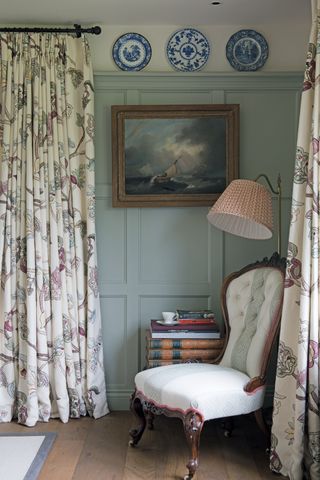 A classic living room with green wall panelled decor, long cream patterned curtains and upholstered chair