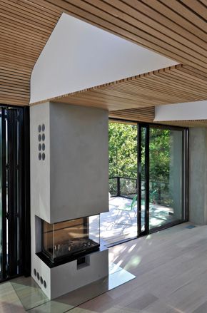 View of a tall, grey fireplace with a glass unit at Trekronekabin in a space with a wood slat covered ceiling, wood flooring and glass doors leading to an outdoor area